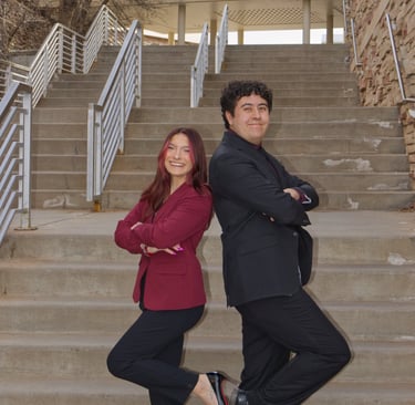 Victoria Quesada-Stoner and Ben Gregg standing back to back with hands folded, and stairs behind.