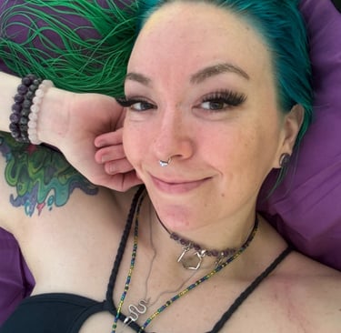 Anointistry creator, woman with green hair, crystal jewelry smiling warmly laying on a purple bed 