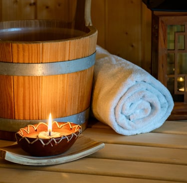 Wellness-Sauna mit Holzeimer, gerolltem weißen Handtuch und stimmungsvoll brennender Kerze.