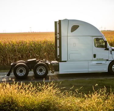 A white semi-truck tractor driving past a sunlit cornfield with wind turbines in the background.
