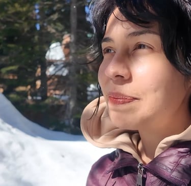 Person standing outdoors in a snowy forest, looking to the side in sunlight with tall trees in the b