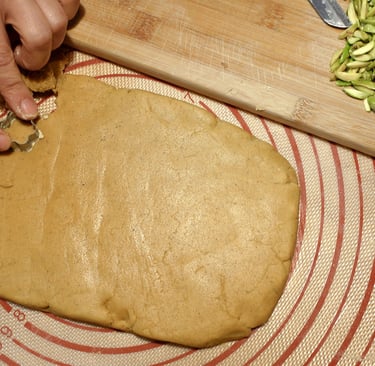 Hands cutting shapes from rolled cookie dough on a silicone mat with pistachios nearby.