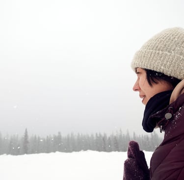 Person in winter jacket and knit hat standing in falling snow, looking across a snowy landscape with