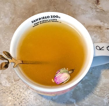 Cup of herbal tea with a small rosebud and spoon inside, placed on a table.