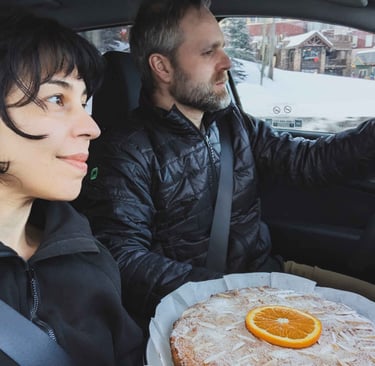 Two people sitting in a car with a homemade almond orange cake resting on a tray between them.