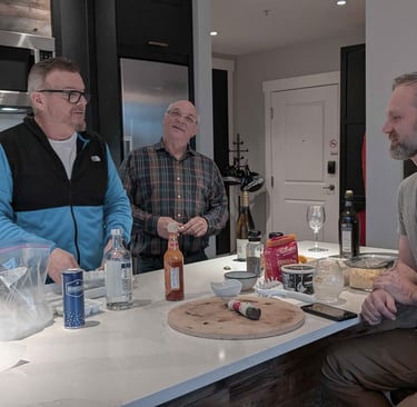 Three men talking around a kitchen island with drinks and food during a casual evening at home.