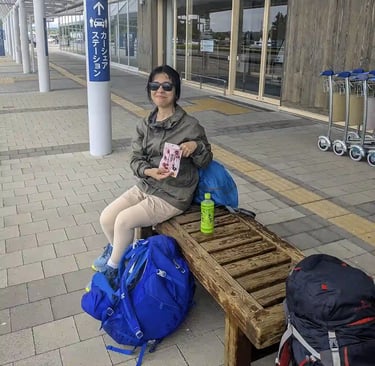 Woman sitting on a wooden bench outside an airport terminal with hiking backpacks and a bottle of green tea