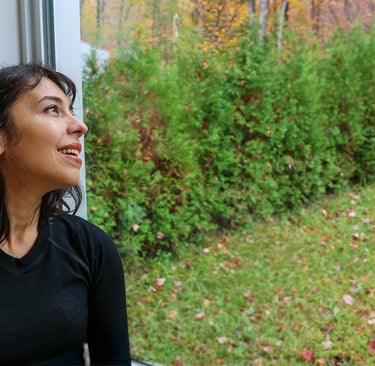 Woman sitting by a window looking outside at evergreen trees and autumn foliage.