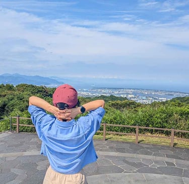 Woman standing at a mountain viewpoint overlooking Shizuoka city, forests, and the Japan Sea coastline.