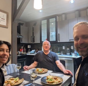 Three people sitting at a dining table in a kitchen, smiling while sharing a home-cooked meal.