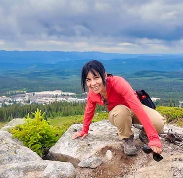 Woman crouching on a rocky mountain viewpoint overlooking Big White and surrounding forests under cloudy skies.