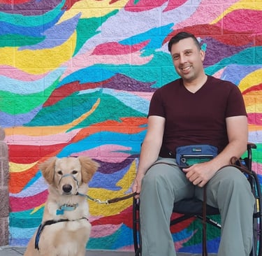A man in a wheelchair with his golden retriever service dog in front of a colorful mural.