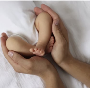 Close-up of a parent's hands gently cradling a newborn baby's tiny feet and legs on white bedding.