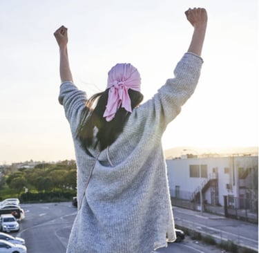 A breast cancer survivor in a pink headscarf raising fists in victory on a sunny balcony.