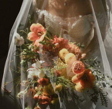 Novia con vestido de encaje bajo velo transparente sosteniendo un ramo en tonos naranja, blanco y verde.