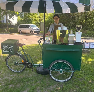 Grünes Kaffeefahrrad mit grün-weißem Schirm und Barista auf einer Wiese.