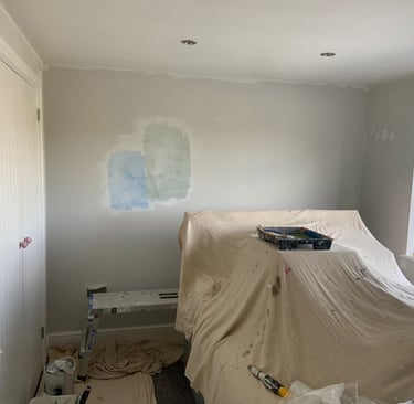 An in-progress bedroom, with sheet covered furniture and evidence of paint-testing on the walls.