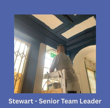 Portrait of Stewart, a senior team leader, on a ladder painting a doorway and facing away.