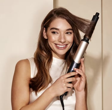 A smiling woman using a ceramic curling wand to create wavy hairstyles at home.