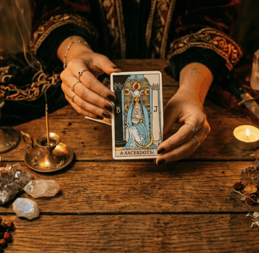 A fortune teller's hands holding The High Priestess tarot card on a wooden table with candles and crystals.