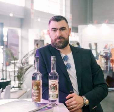Bearded man in a navy blazer presenting bottles of Suspiro d’Alma artisanal mezcal at a trade show.