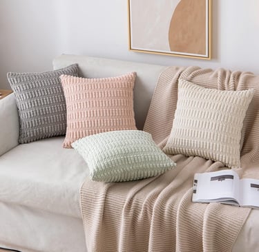 Textured decorative throw pillows in pastel colors arranged on a white sofa with a beige knit blanket.