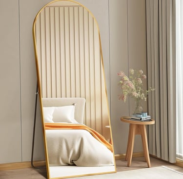 Full length arched floor mirror with gold frame leaning against a bedroom wall next to a wooden side table.