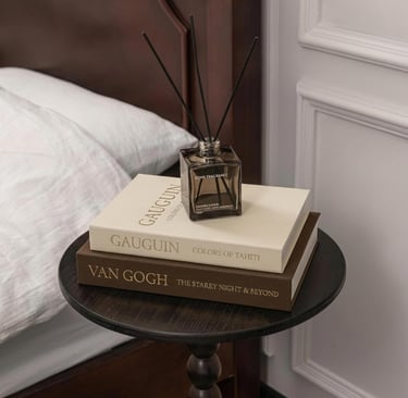 A reed diffuser sits on top of Van Gogh and Gauguin art books on a dark wood bedside table.