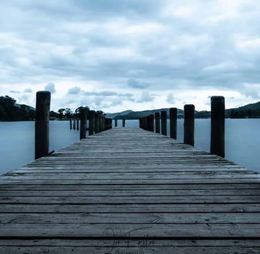 A long wooden pier stretching into a calm blue lake under a cloudy sky with mountain silhouettes.