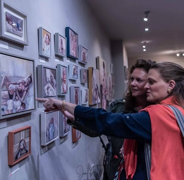 Two women looking at a gallery wall of framed contemporary portrait paintings in an art exhibition.