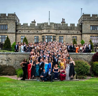 A large group of graduates in formal attire posing for a photo in front of a historic castle venue.