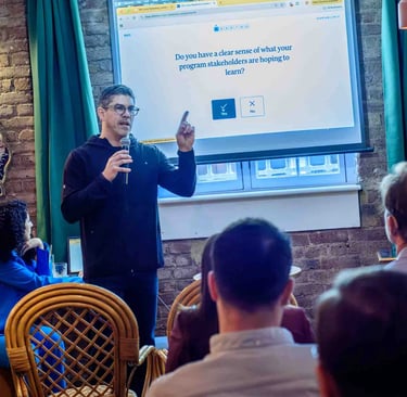 A man presents a question about program stakeholders to an audience, using a projector in a dimly lit room during a business