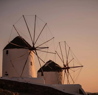 Traditional white Mykonos windmills at sunset, a scenic landmark in Greece with a golden sky.