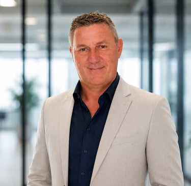 Professional portrait of a middle-aged male executive in a light gray suit jacket inside a modern office.