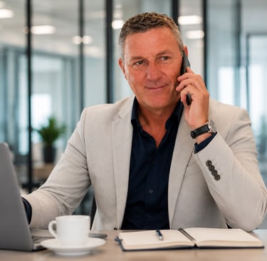 Professional businessman in a suit talking on a mobile phone while working on a laptop in a modern office.
