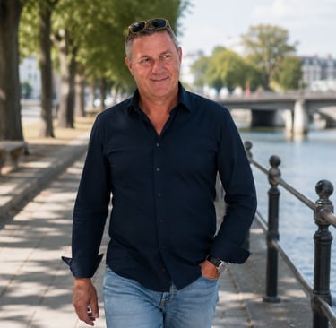 A middle-aged man in a navy shirt and jeans walking along a riverside path with sunglasses.