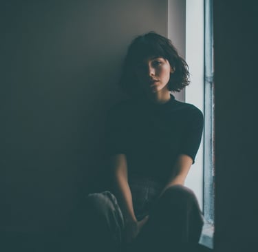 Moody portrait of a woman with short hair sitting by a window in a dimly lit room.