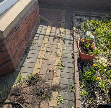 A brick patio walkway with scattered garden soil and weeds next to a planter box.