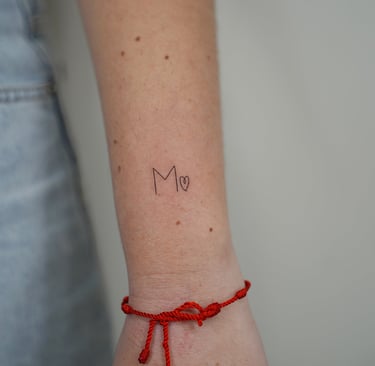 Tatuaje fineline en Madrid de inicial M con corazón pequeño en trazo fino en la muñeca