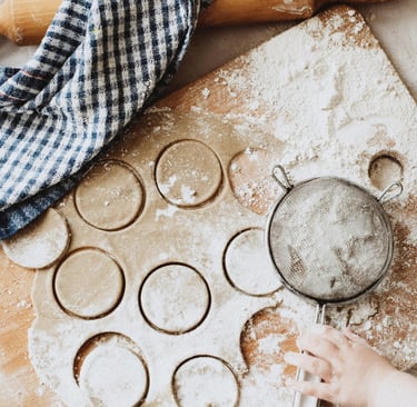A child's hand using a flour sifter on cookie dough cut into circles on a wooden baking board.