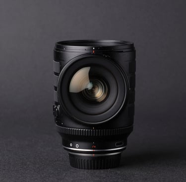A minimalist studio shot of a professional camera lens on a dark surface. The focus is on the subtle off-white light reflecting off the glass elements. Clean, sophisticated composition set in a North American / US photography studio.
