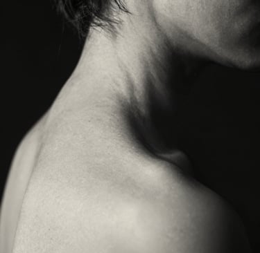 A fine art monochrome photography study of human form, focusing on the curve of a neck and shoulder in dramatic low-light. Deep black shadows and soft off-white highlights create a sense of depth. North American / US artistic style.