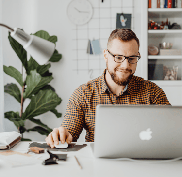 Smiling man with glasses working on a laptop at a bright home office desk.