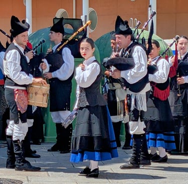 A group of musicians in traditional black and white folk attire play bagpipes and drums in an outdoor plaza in Cuntis.