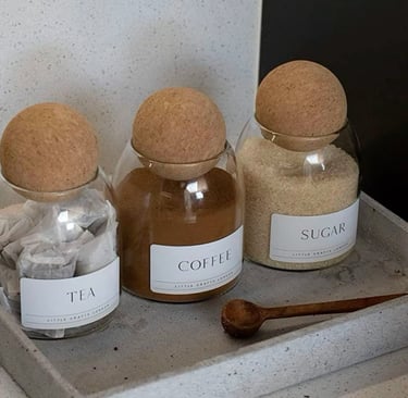 Minimalist glass storage jars for tea, coffee, and sugar with cork ball lids on a concrete tray.