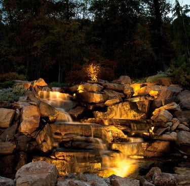 Beautiful, illuminated rock waterfall with lush trees, creating a serene and natural garden landscape at dusk.