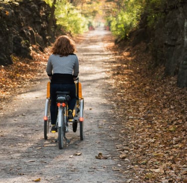 A woman with shoulder length red hair rides a three-wheeled wheelchair bike away from the camera.
