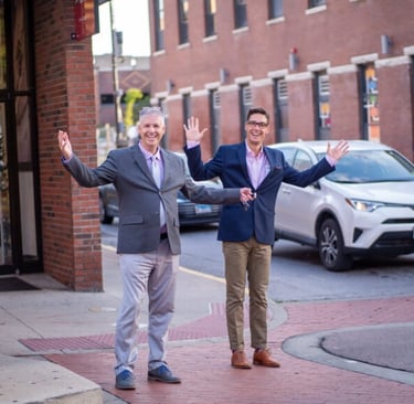 Lawrence and another man standing at a crosswalk with arms up smiling at the camera