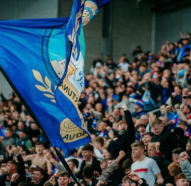 Linfield-Fan in blauem Trikot vor dem Windsor Park in Belfast, mit Blick auf die moderne Stadionfassade und die Tribünen im H
