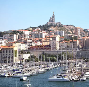 Blick auf Marseille vom Meer aus mit Fischerbooten im Vordergrund, die Altstadt und der Hafen sichtbar, im Hintergrund die Ba